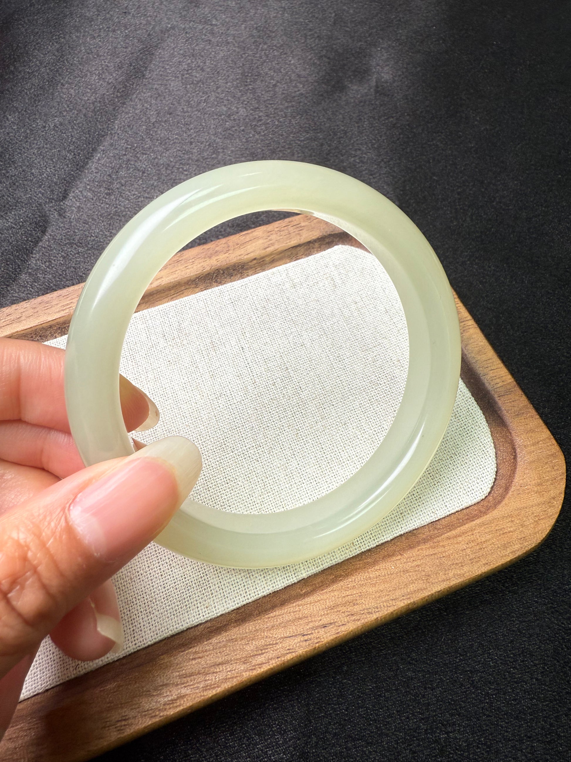 Hand holding a light green hetian jade Bangle on a wooden tray with a dark background