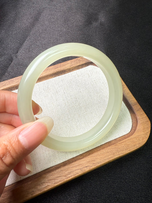 Hand holding a light green hetian jade Bangle on a wooden tray with a dark background